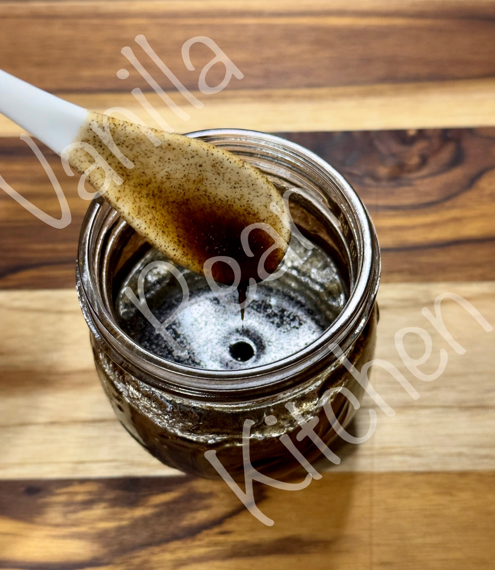 Load video: Video image of a clear jar full of thick brown viscous whole bean vanilla paste being stirred with white ceramic spoon.